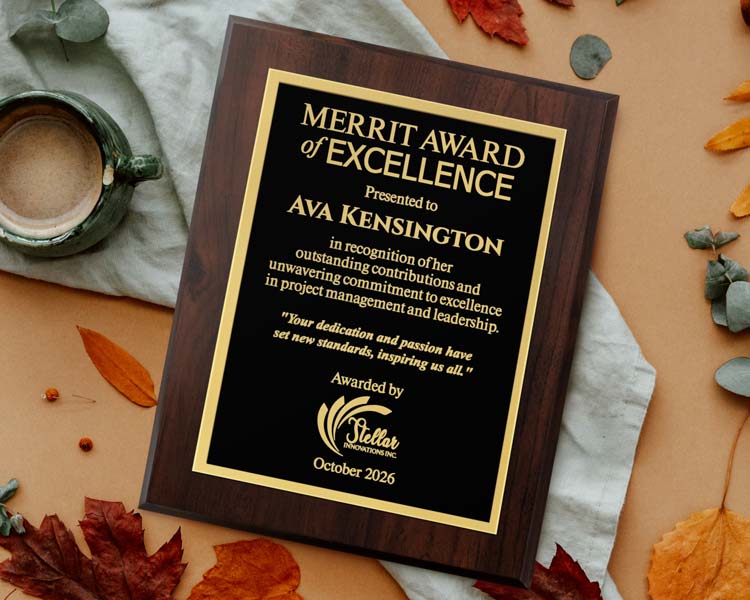 A laser engraved plaque with gold engraving on a cherry wood board, laying on a table surrounded by fall themed leaves and a mug. Engraved on the front is a merit award for Ava Kensington.