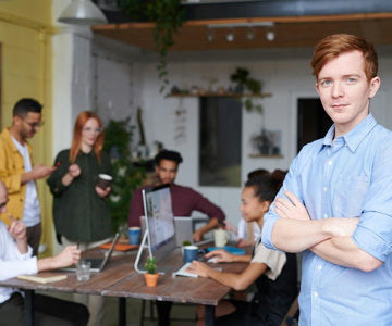 Happy Employee Crossing Arms Near Coworkers Around An Office Desk Working On Computers And Drinking Coffee