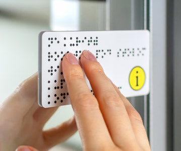 Pair Of Fingers Brushing Across A White Label Featuring Braille And Information Logo And Mounted Onto A Mirror