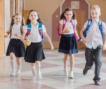 Three Little Girls And One Little Boy In School Uniforms Carrying Backpacks And Apples Through A School Hallway
