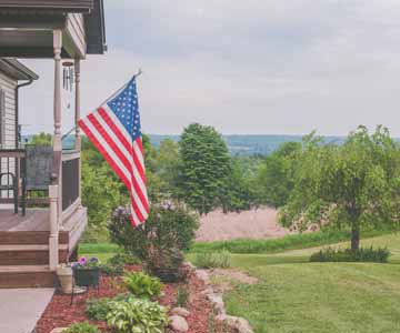 An American Flag in a flag stand on the front porch of a country house.