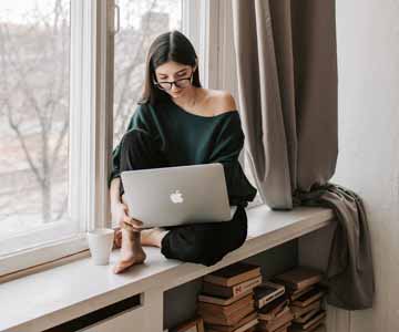 A young woman sitting by a window, working on her laptop computer.