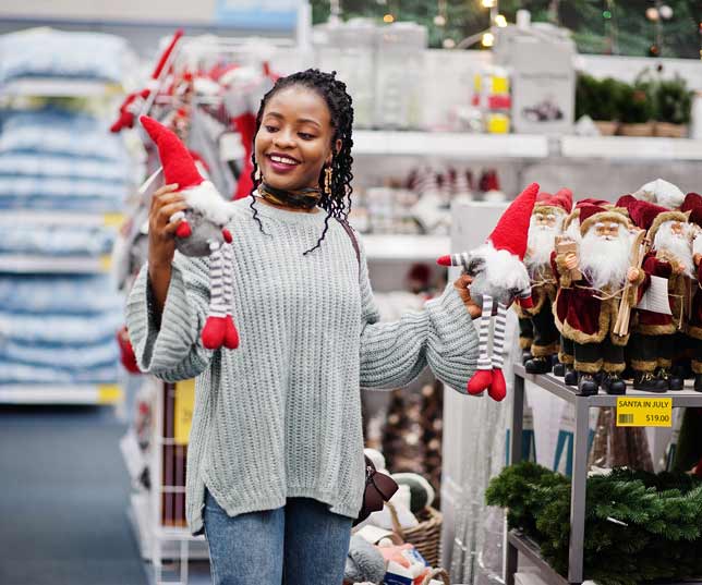 Happy Woman Shopping In Store And Holding Up Two Christmas Gnome Dolls Beside Aisle Of Santa Claus Dolls And Decor