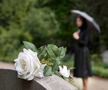 A woman in black stands in the background, visiting a grave with a white rose placed gently on top.