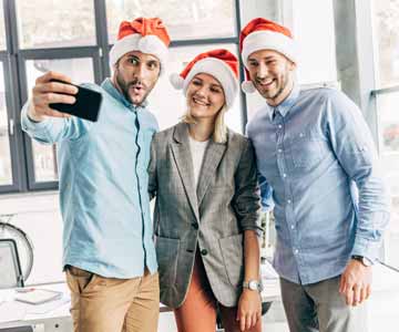 Three coworkers taking a picture at the office Christmas party wearing Santa hats. 