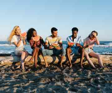3 women, 2 men eating watermelon on a log by the seashore