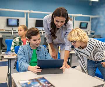 Woman Teacher In Classroom Smiling And Leaning Between Two Small Students At Desks