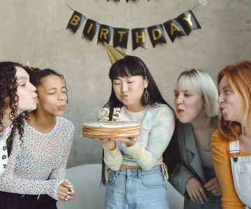 A group of five women blowing out a candle on a birthday cake with a 25 shaped candle