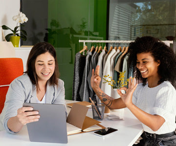 Two Happy Women Working At A Small Business Work Table With Laptop And Phone And Notepad In Front Of Clothes Rack