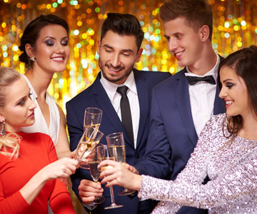 Group Of Happy Coworkers Dressed In Formal Party Attire Indoors Near A Gold Party Curtain And Making A Toast With Glasses