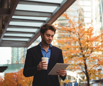 Happy Man Wearing A Business Suit And Walking Outdoors By Trees With Fall Orange Leaves And Holding A Coffee Cup