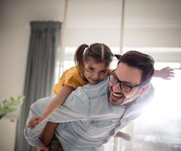 Dad In Glasses Giving Young Daughter A Piggyback Ride In Living Room