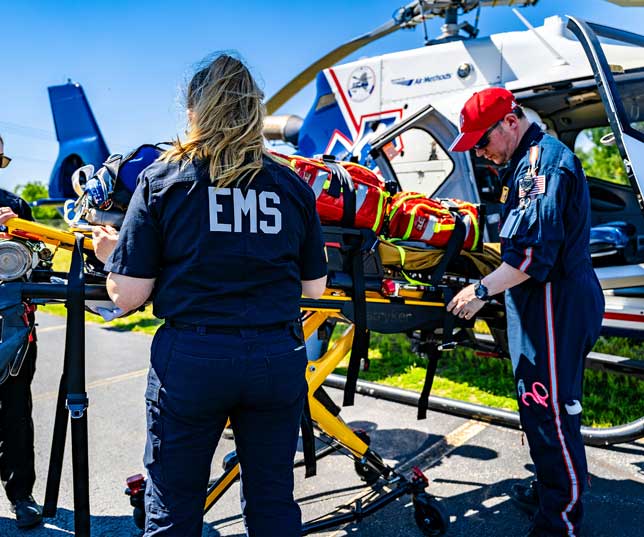 Two EMTs Wearing Uniforms And Helping Victim Onto A Stretcher And Moving Them Into A Helicopter Outdoors In The Afternoon