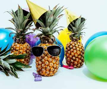 Three pineapples, one wearing sunglasses, surrounded by balloons and party favors.
