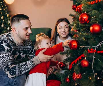 Two Young Parents With Little Child Decorating Christmas Tree In Living Room With A New Ornament