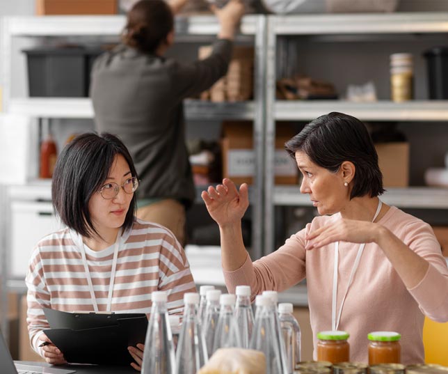 Two Women At Counter With Boss Explaining Something To Employee While A Stocker Works On Shelves In Background