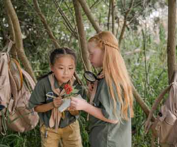 Two Summer Camp Attendees Using Magnifying Glass To Look At Insects.