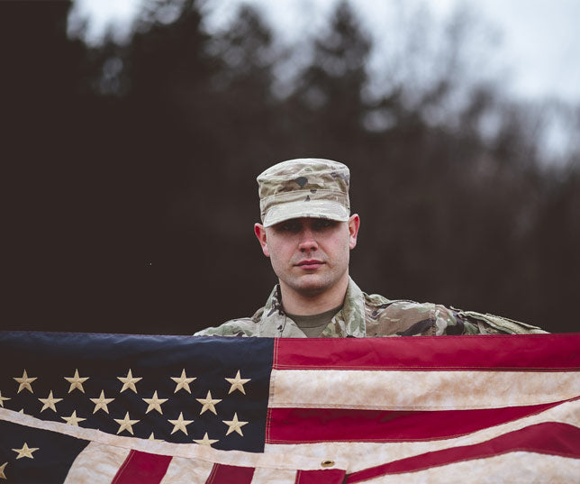 United States Soldier In Full Military Uniform Standing Outdoors Near Edge Of Woods And Holding Up The American Flag