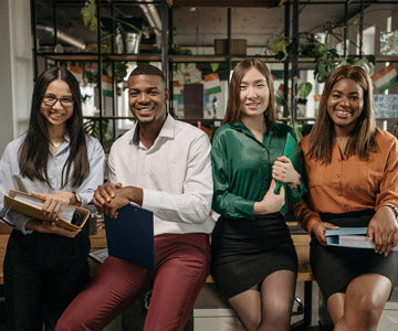 Four Happy Coworkers In Business Attire And Holding Folders And Sitting Beside Each Other In Office Nearby Shelves