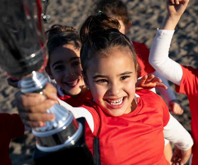 Group Of Happy Little Girls Outdoors On Beach Wearing Red Uniforms With One Girl Holding Up A Silver Trophy Cup