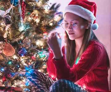 Woman Dressed In Santa Outfit Sitting On The Floor Beside Brightly Lit Christmas Tree And Decorating It With Lights