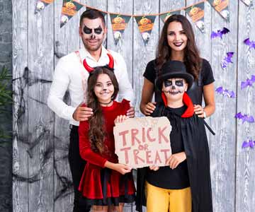 Parents With Two Little Kids Wearing Halloween Costumes In Backyard In Front Of Gray Fence With Halloween Decorations