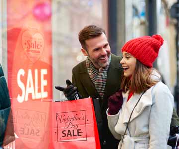 Couple On Sidewalk Looking Into Business Window With Bright Red Sign Featuring Valentine’s Day Sale Details