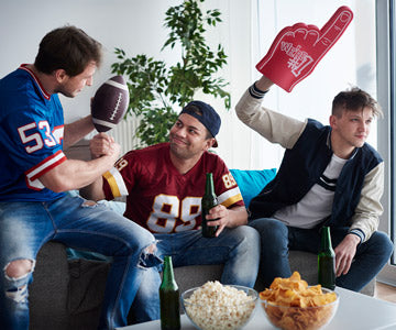 Three Men Wearing Football Jerseys Sitting On Living Room Couch Near Coffee Table With Beer Bottle And Snack Bowls