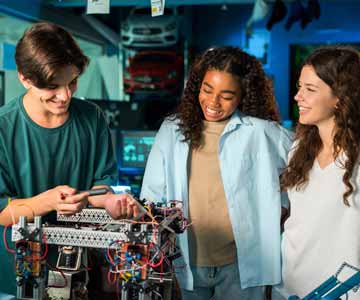 Three Teenage Students Dressed In Casual Attire And Smiling As They Work In A Computer Lab