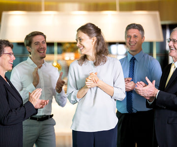 Group Of Coworkers In A Brightly Lit Office Applauding For A Happy Businesswoman In Front Of Shelving