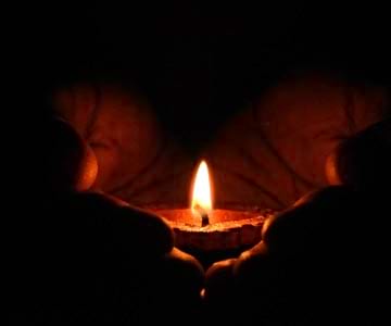 Dark hands gently hold a small glowing candle in the palm.
