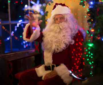 Santa sits on a chair beside Christmas garland lit with twinkling lights, ready for the season.