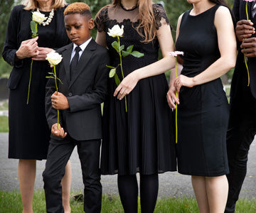 Little Boy Outside At Cemetery With Group Of Adults Dressed In Black And All Carrying A Single White Flower Each