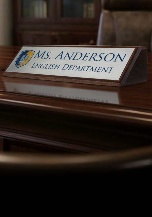 A walnut desk wedge with a silver aluminum metal plate on the front reading ‘MS. ANDERSON ENGLISH DEPARTMENT’ in blue text, placed on a polished wooden desk