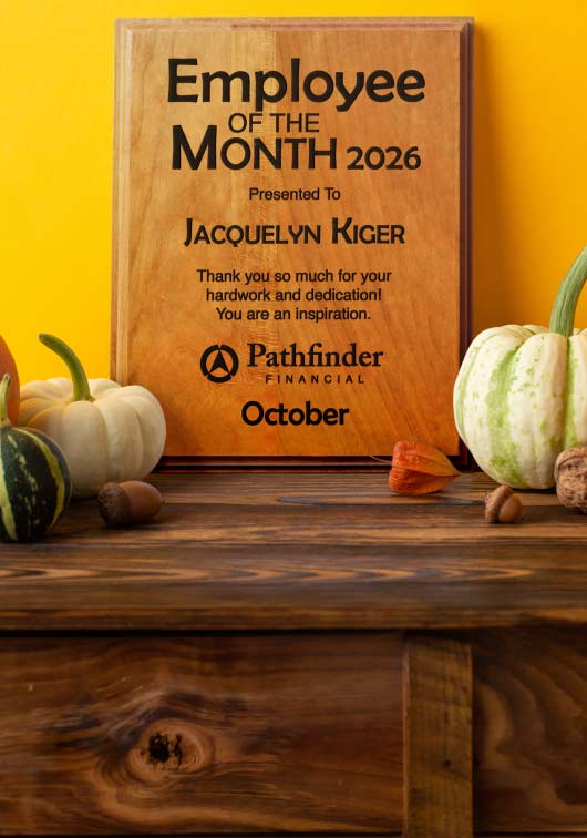A solid cherry wooden plaque with engraved text about an Employee of the Month award presented to Jacquelyn Kiger. Displayed on a rustic wooden table with small pumpkins against a yellow wall.