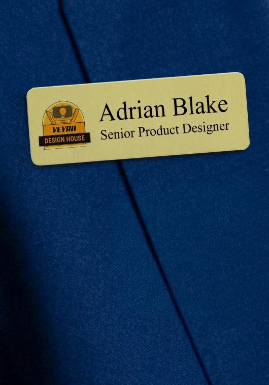 A gold aluminum metal name tag with rounded corners and printed black text  reading ‘Adrian Blake’ with a title and a logo beside the name. Placed on a textured royal blue fabric background.