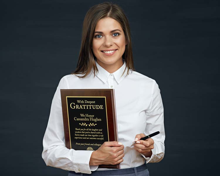 A smiling woman in a white button-down shirt holds a personalized plaque with a wooden board that reads ‘With Deepest Gratitude’ in gold engraved text, against a dark charcoal background.