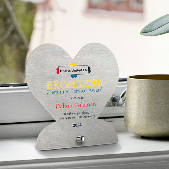 A heart-shaped stainless steel award on a windowsill, featuring colorful printed text for a customer service award. A gold cup sits on the sill next to it, with a blurred outdoor background.