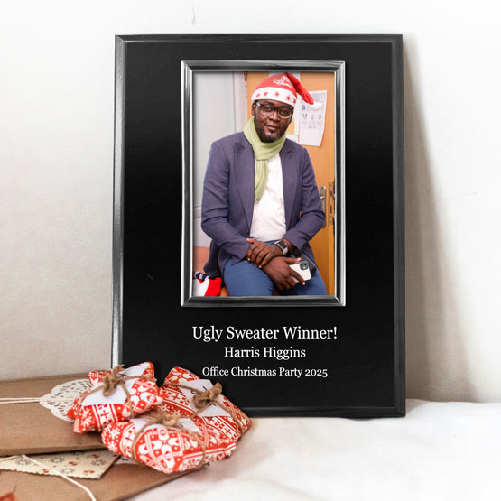 A personalized photo frame on a black board with silver frame next to wrapped Christmas gifts. The frame contains a picture of a smiling man sitting down, with text printed on the board that reads ‘Ugly Sweater Winner! Harris Higgins Office Christmas Party 2025’.