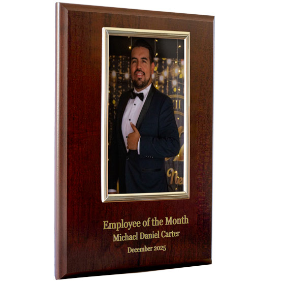 A cherry wood board featuring a gold picture frame that contains a photo of a man holding a present. Below the picture frame is gold printed text about an Employee of the Month award for Michael Daniel Carter, set against a white background.