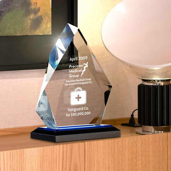 A faceted acrylic award on a black base, displayed on a wooden console table next to a lamp in an indoor setting. The acrylic features custom laser engraved text and a shield logo.