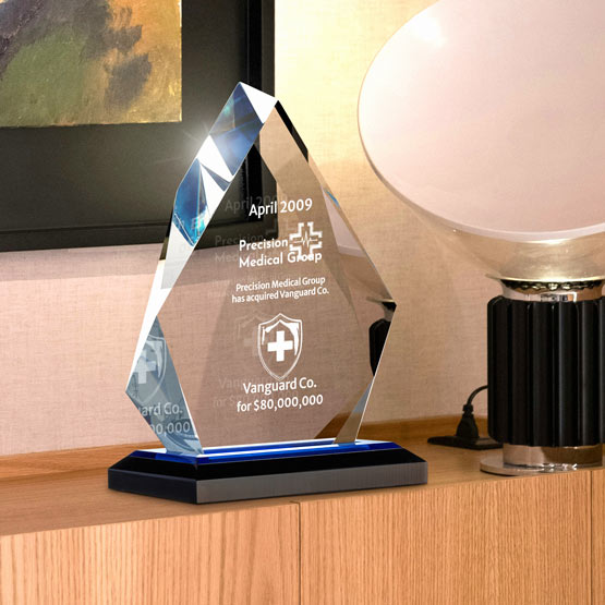 A faceted acrylic award on a black base, displayed on a wooden console table next to a lamp in an indoor setting. The acrylic features custom laser engraved text and a shield logo.