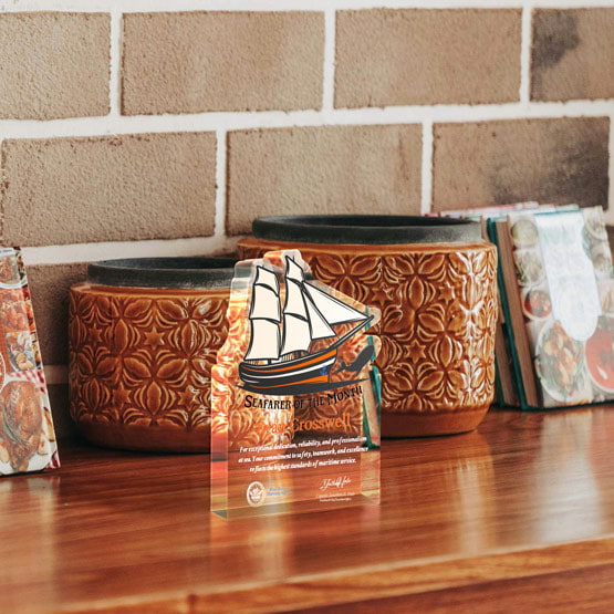 Five Inch Sailboat Shaped Acrylic Award For Seafarer Printed With Ship Logo And Message Freestanding On Wood Table With Pots