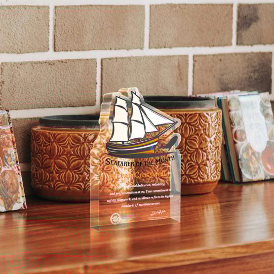 Six Inch Sailboat Shaped Acrylic Award For Seafarer Printed With Ship Logo And Message Freestanding On Wood Table With Pots