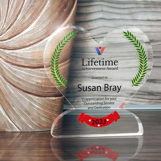 A clear acrylic heart-shaped award with green printed laurel wreaths and a colorful logo presenting a ‘Lifetime Achievement Award’ to ‘Susan Bray’, displayed on a reflective wooden surface beside a textured vase.