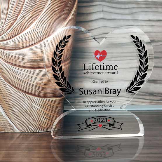 A clear acrylic heart-shaped award with black printed laurel wreaths and a red heart logo presenting a ‘Lifetime Achievement Award’ to ‘Susan Bray’, displayed on a reflective wooden surface beside a textured vase.