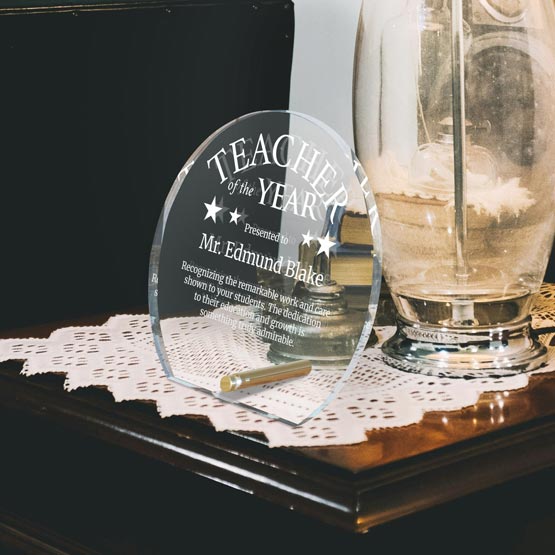 A clear acrylic Teacher of the Year award plaque engraved with the text 'Presented to Mr. Edmund Blake' and a gold-colored pen resting inside its base, displayed on a white lace doily on a dark wooden table next to a glass jar.