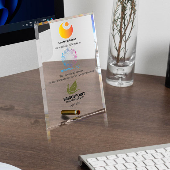 A clear acrylic economy award featuring color printed company logos and text about a 50% stake acquired in ‘Beacon, Co. by Summit Industrial’, standing on a wooden desk next to a keyboard and computer monitor.