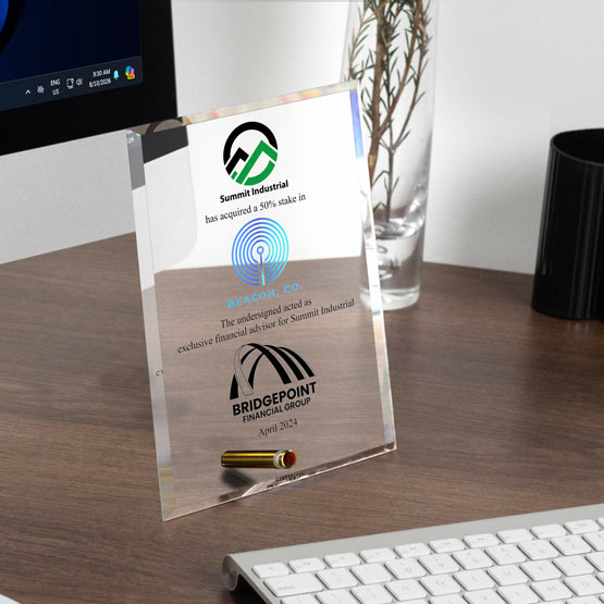 A clear acrylic economy award featuring color printed company logos and text about a 50% stake acquired in ‘Beacon, Co. by Summit Industrial’, standing on a wooden desk next to a keyboard and computer monitor.