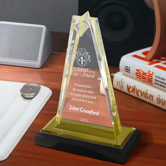A personalized acrylic star-shaped award with a black base and customized engraved text, displayed on a wooden desk next to a speaker, mouse pad, and books.